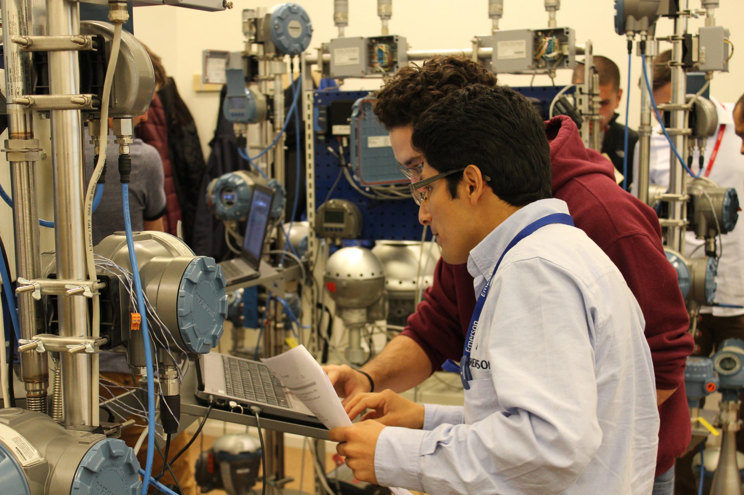 Dos ingenieros trabajan en un laboratorio de automatización industrial, revisando documentación y datos en una computadora portátil conectada a equipos de control de procesos